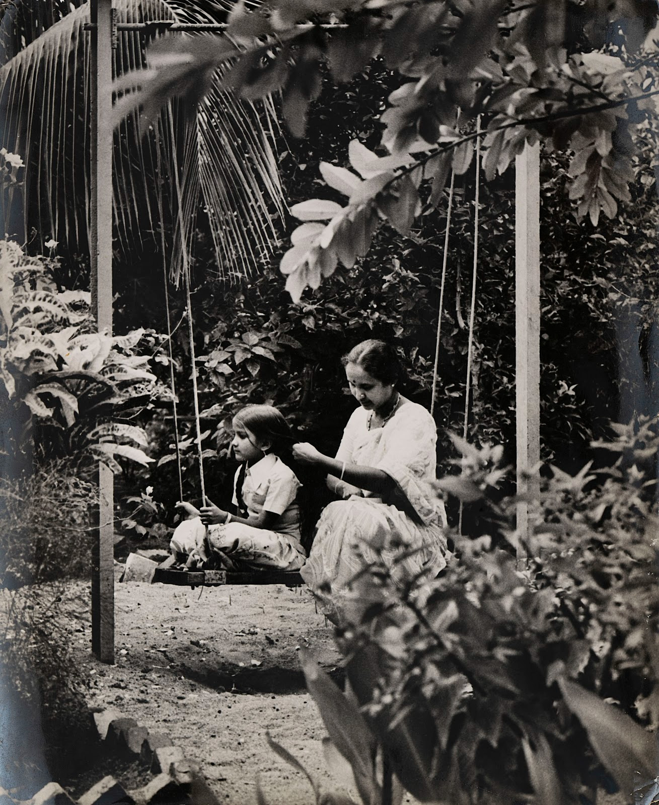 In this black-and-white image from Gawribidanur, Satyan captures an adult woman and child in the ordinary, intimate act of hair braiding, while seated on a swing in a garden.