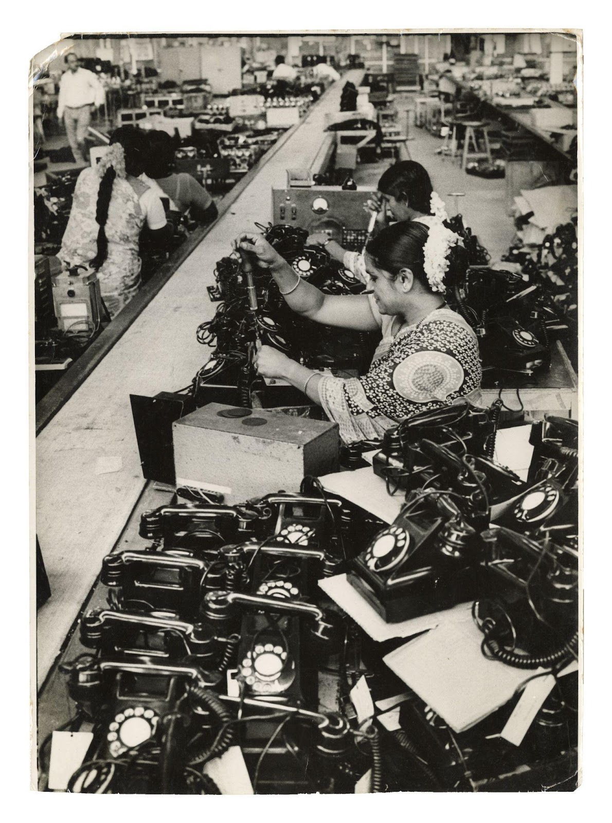 In this black and white photograph we see women working in a telephone factory. There are four women working in the photograph. They are dressed in patterned saris, simple jewellery and flowers in their hair. In the top-left corner, there’s a smaller figure of a formally dressed man walking past, suggesting routine surveillance by a senior.