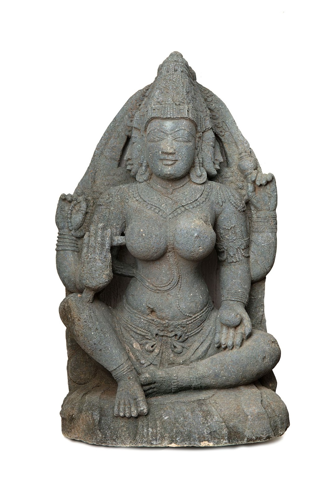 A grey steatite schist sculpture of a female with three faces and four hands. She is seated with her eyes closed and each of her three heads has a crown on it.