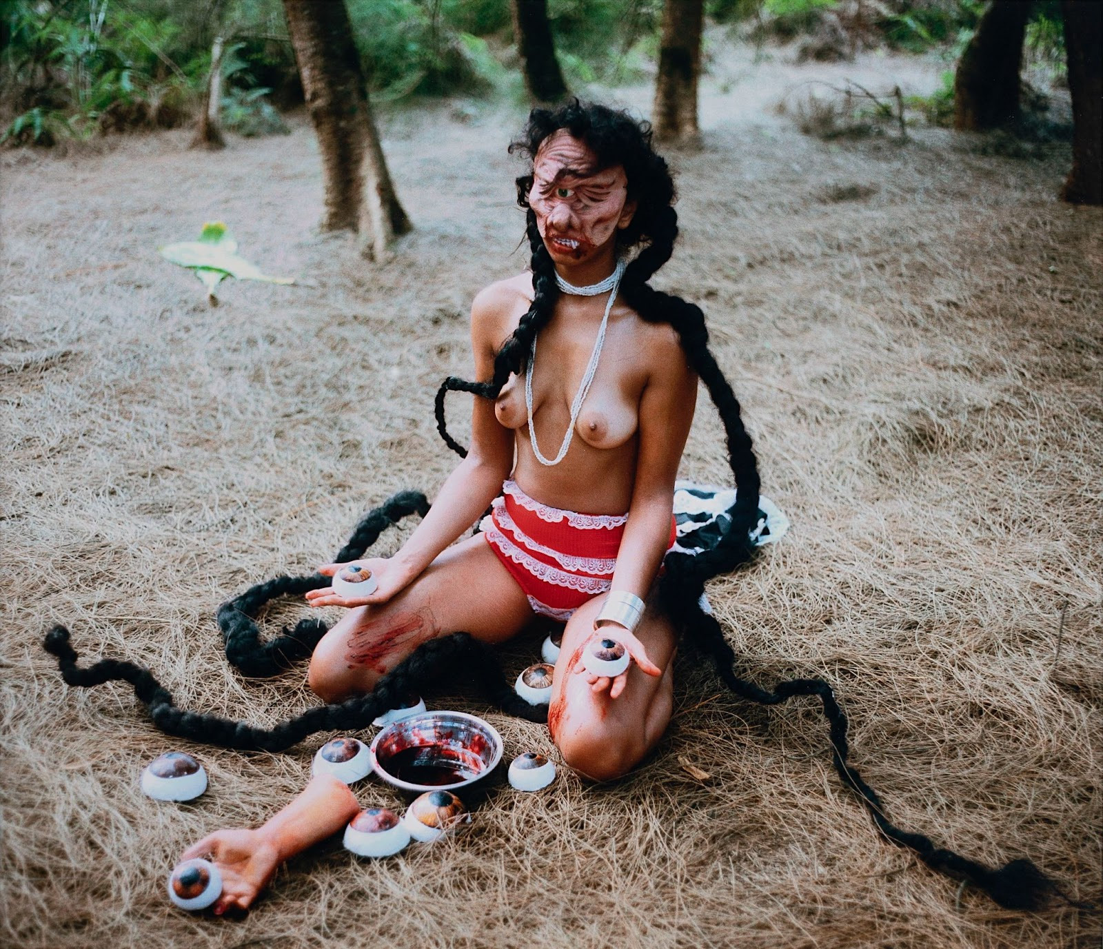 This is a staged photograph is part 2 of three works. It shows the artist as a bare-breasted subject kneeling and performing a blood ritual while wearing a mask of a fierce distorted face.